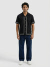 Man wearing a black short-sleeve shirt with white patterns and dark blue jeans on a white background