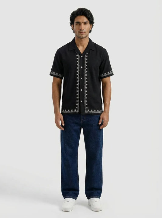 Man wearing a black short-sleeve shirt with white patterns and dark blue jeans on a white background