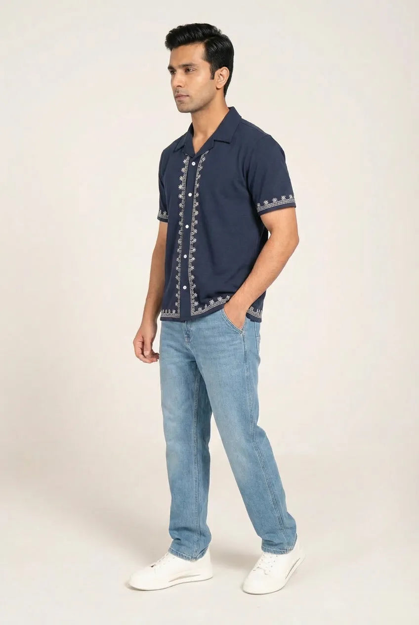 Man wearing a navy blue embroidered shirt and light blue jeans on a white background