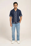 Man wearing a navy blue shirt with white embroidery and light blue jeans on a beige background