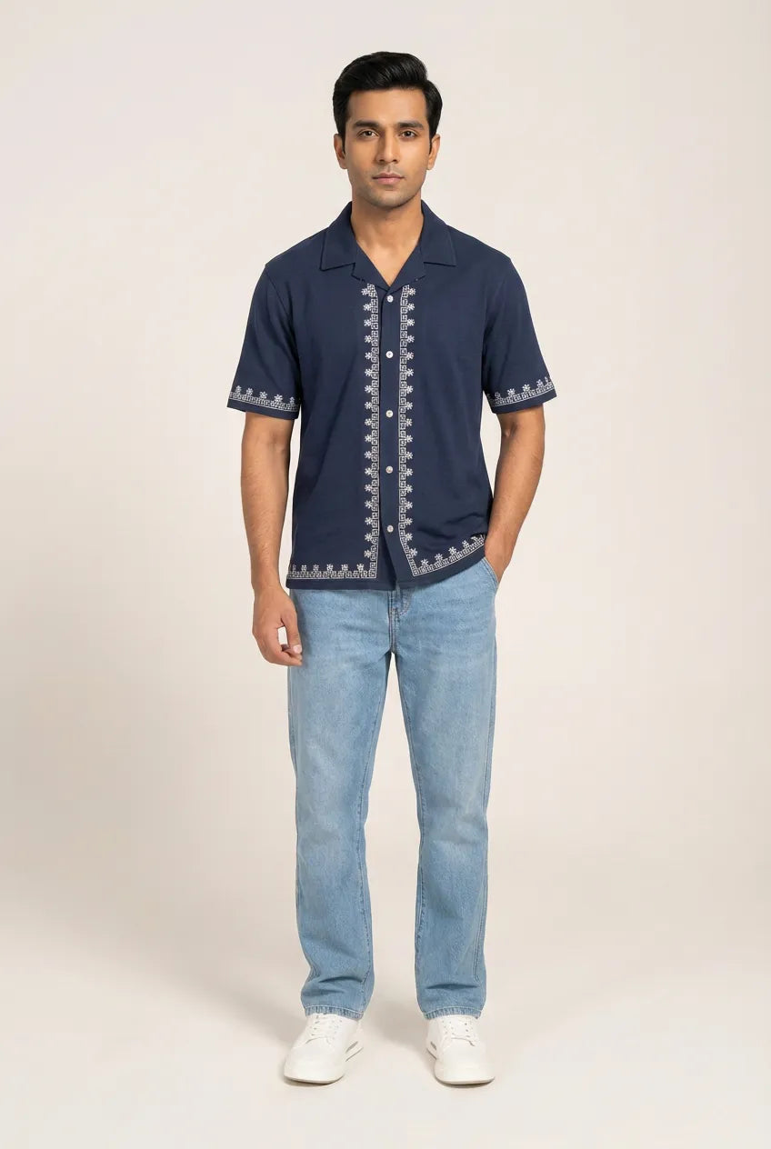 Man wearing a navy blue shirt with white embroidery and light blue jeans on a beige background
