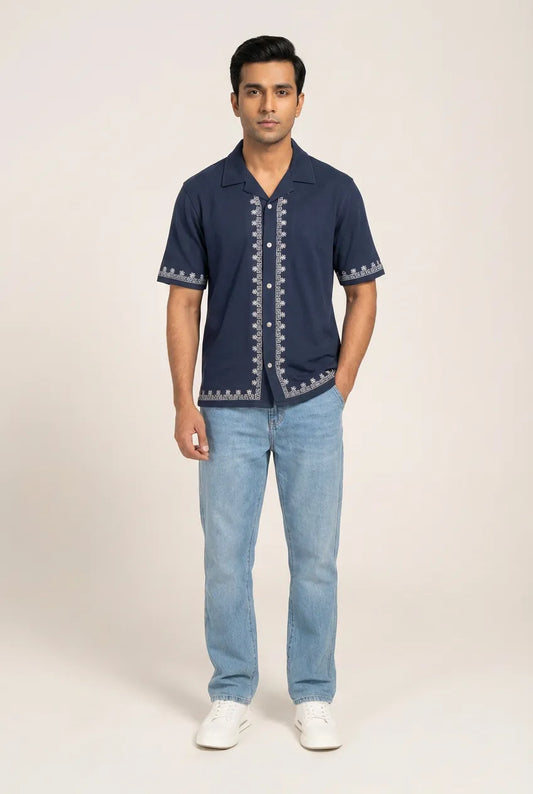 Man wearing a navy blue shirt with white embroidery and light blue jeans on a beige background