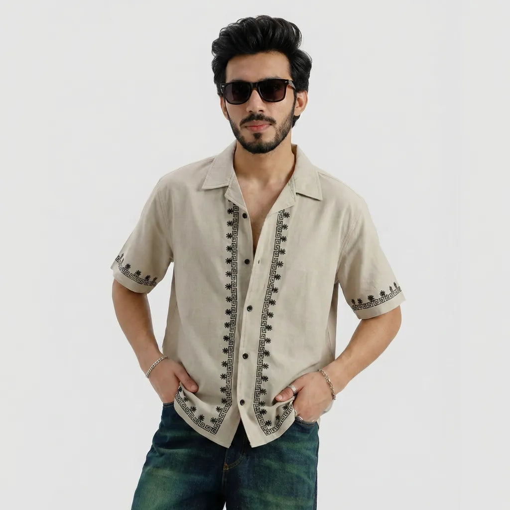 Man wearing a beige short-sleeve shirt with black embroidery and sunglasses on a light gray background